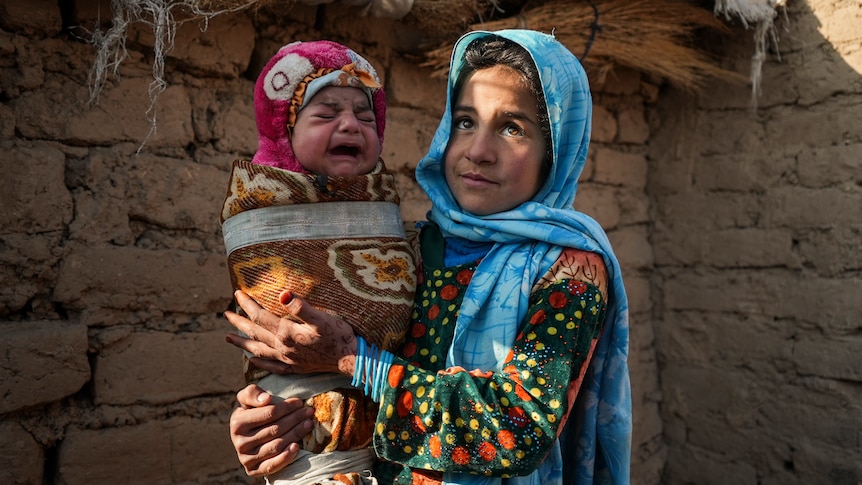 A girl in colourful Afghan garb holds a crying baby. 