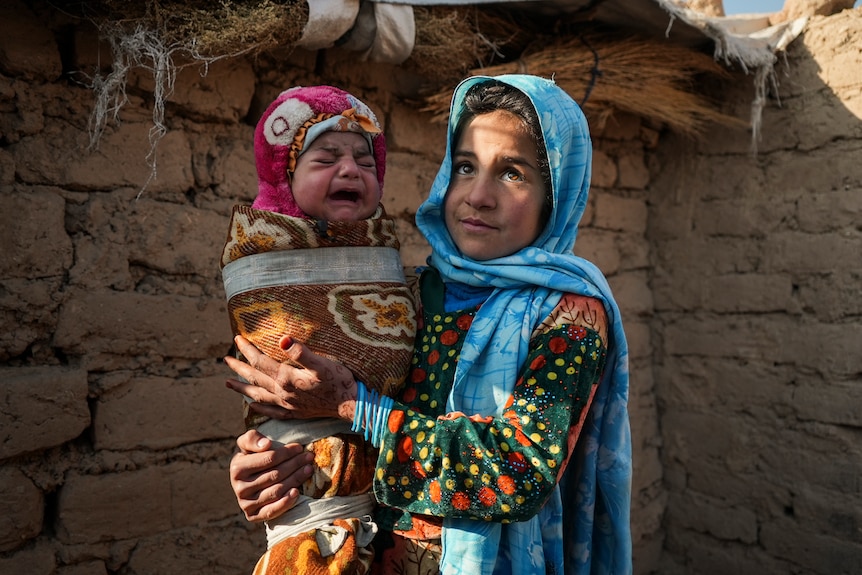 A girl in colourful Afghan garb holds a crying baby. 