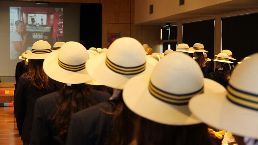A crowd of students sit in a hall with white wide brimmed school hats on.