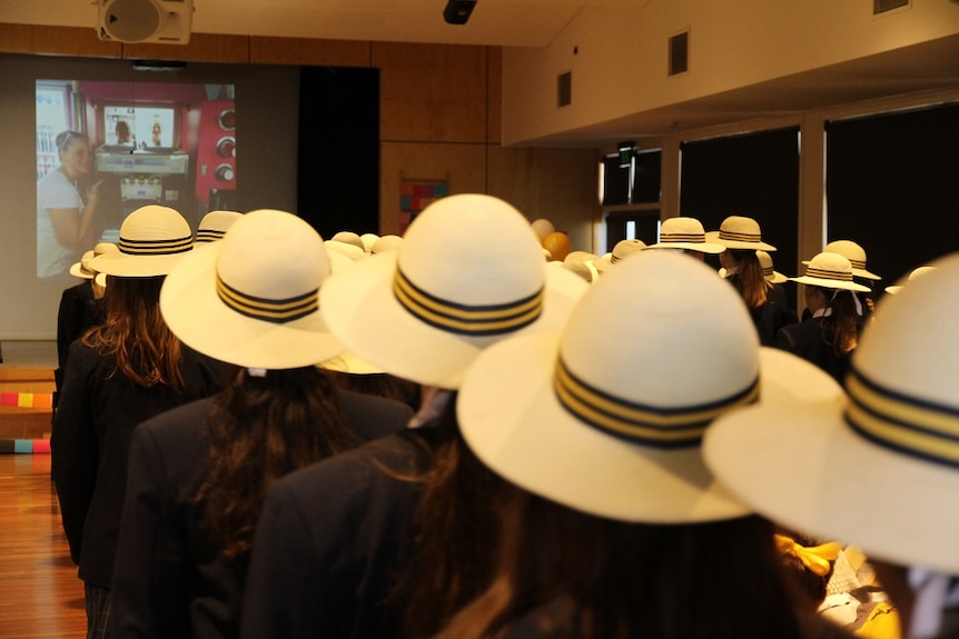 A crowd of students sit in a hall with white wide brimmed school hats on.