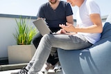 Two men on a bright rooftop looking at a grey laptop.