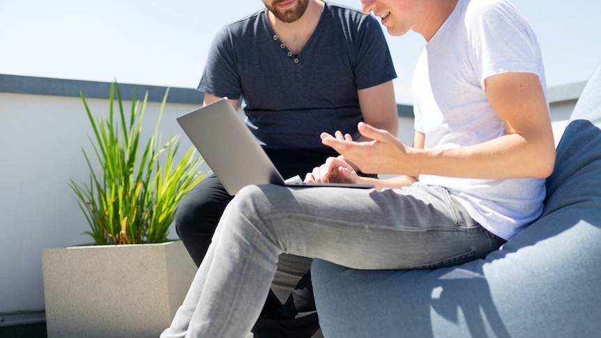 Two men on a bright rooftop looking at a grey laptop.
