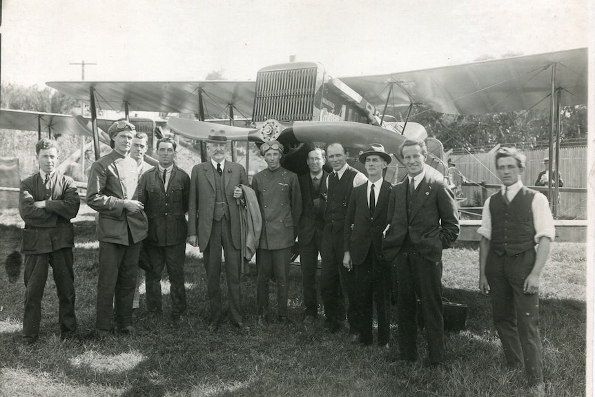 A historic photo of a group of men standing in front of an old aeroplane.