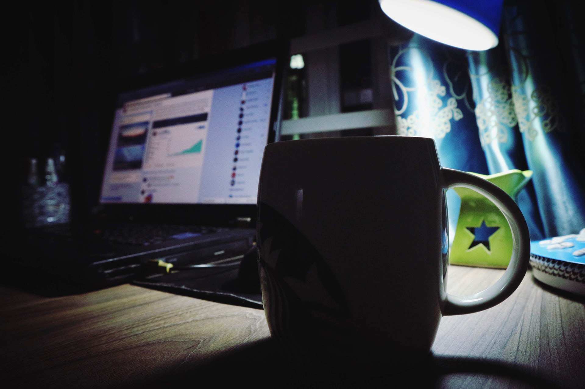A ceramic coffee mug sits on a desk near a laptop and a desk lamp in a dark room.