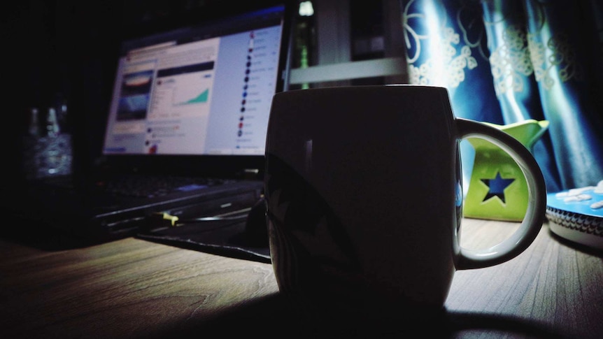 A ceramic coffee mug sits on a desk near a laptop and a desk lamp in a dark room.