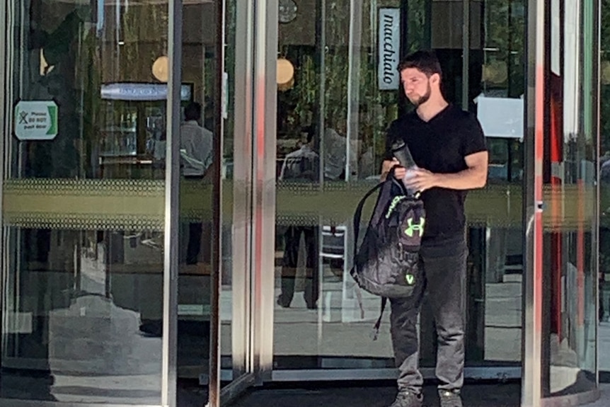 A man with a backpack stands outside large glass doors.