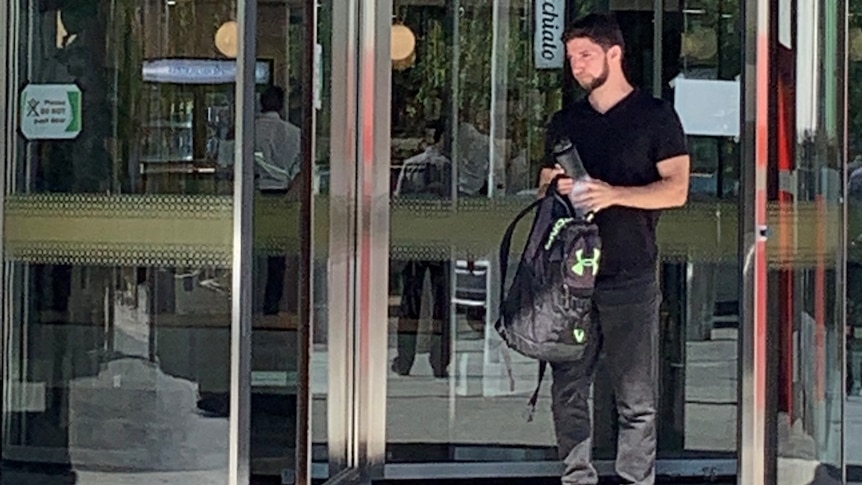 A man with a backpack stands outside large glass doors.