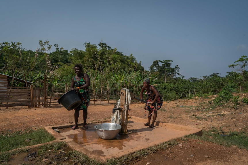 One woman works the pump on a traditional water pump while another cleans out a large bucket