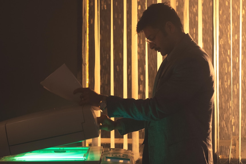 A man stands at a photocopier, mostly in the dark but lit by a window and the glow of the machine