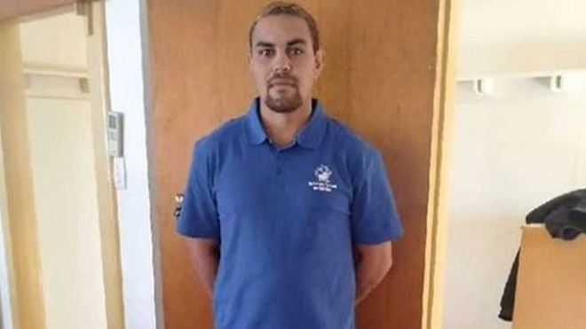 An Indigenous man in a blue polo shirt and black trousers stands with his hands behind his back.