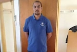 An Indigenous man in a blue polo shirt and black trousers stands with his hands behind his back.