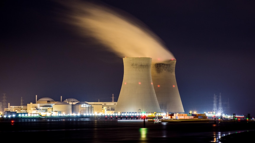 A long exposure shows a nuclear plant illuminated at night, with a white cloud of steam emerging from the top of two reactors. 