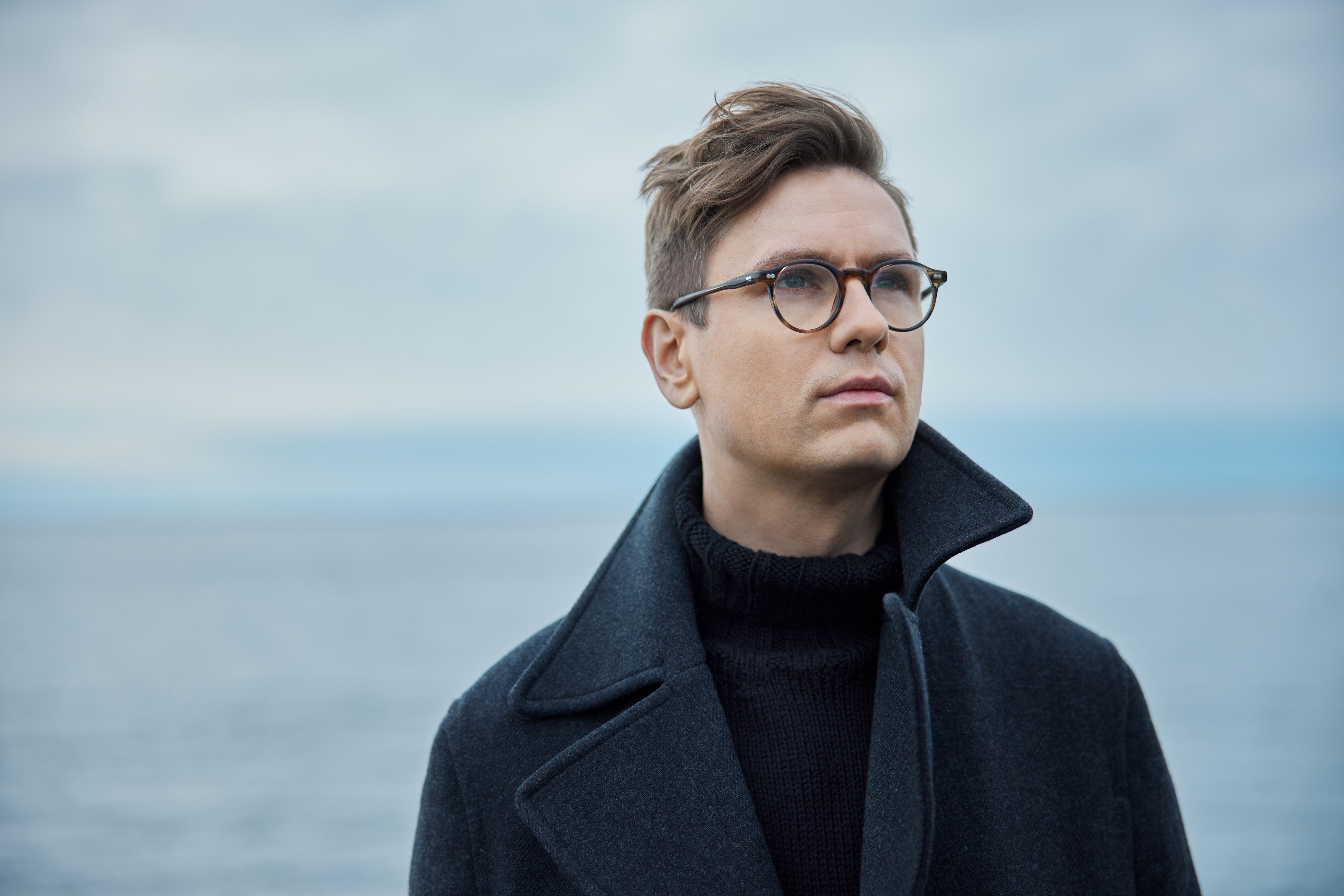 A man wearing glasses looks up into the distance. He wears a dark coat and stands with his back to the sea.