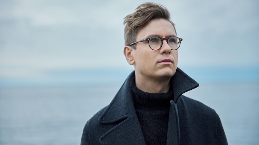 A man wearing glasses looks up into the distance. He wears a dark coat and stands with his back to the sea.