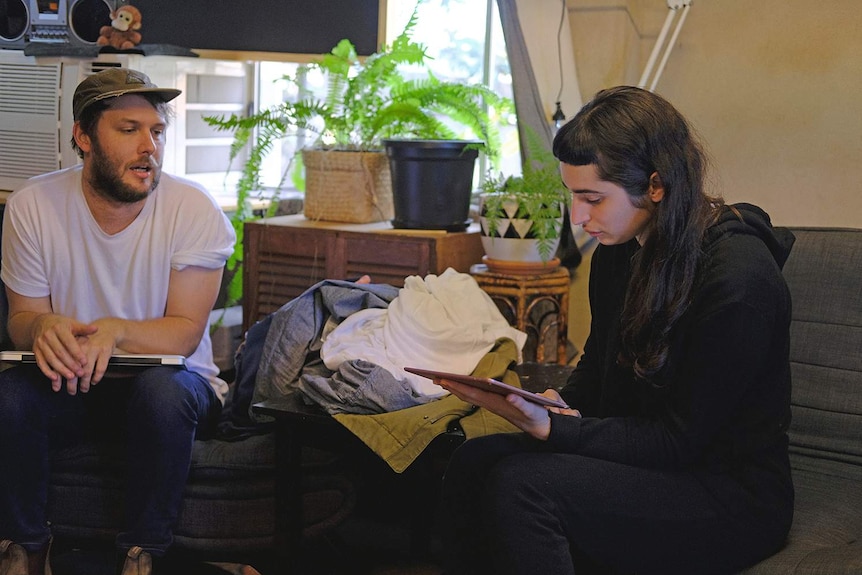 The two young people sit in a messy office space, Macdonald reading from a pink iPad.