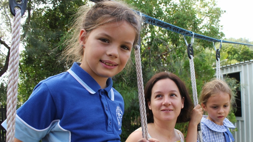 Two young girls in school uniform, on a swing set. Their mother is between them