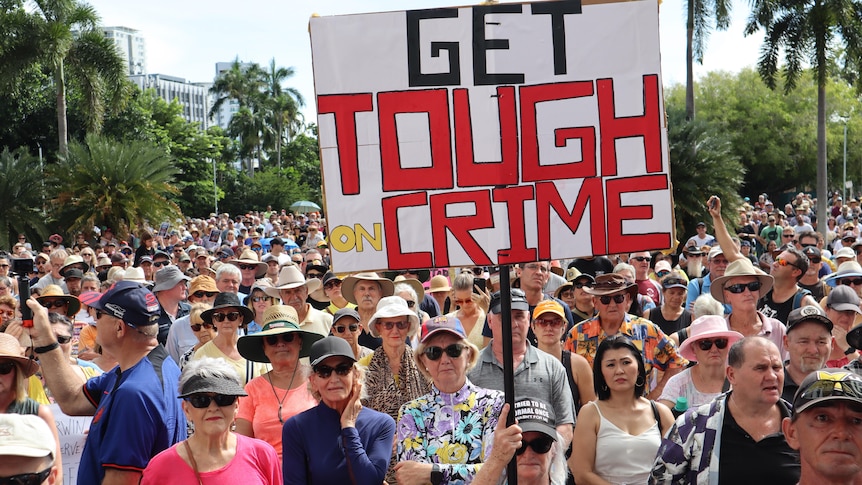 A large crowd of people gathered in a town square, with a large prominent sign reading 'Get Tough on Crime'