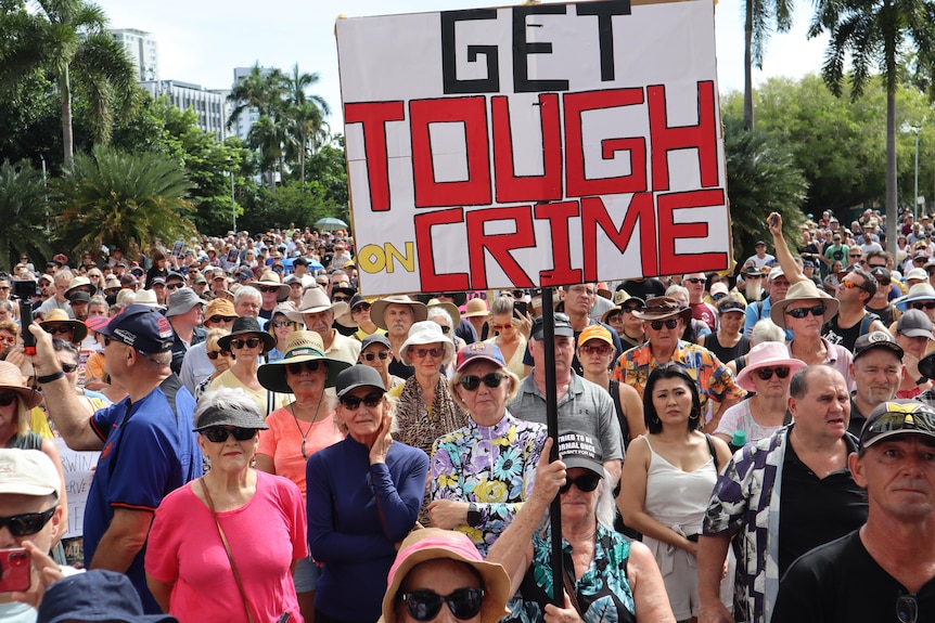 A large crowd of people gathered in a town square, with a large prominent sign reading 'Get Tough on Crime'