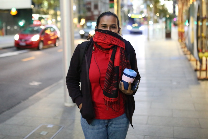 A woman standing on a sidewalk in the Perth CBD holding a coffee cup and with a red scarf around her face poses for a photo.