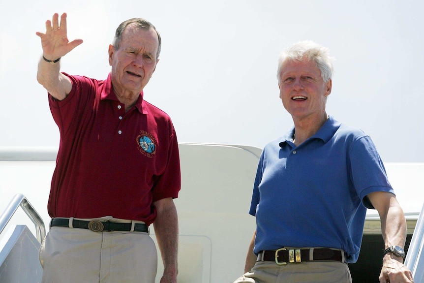George H W Bush waves while standing beside Bill Clinton while exiting a plane.