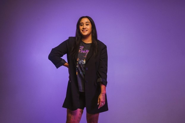 A woman wearing a black jacket poses in front of a purple background, one hand on her hip.