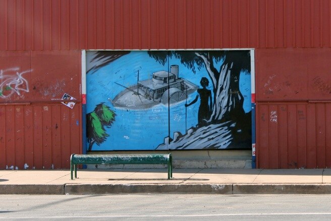 A mural on a red wall depicting a paddle steamer boat floating down a blue river watched over by aboriginal people.