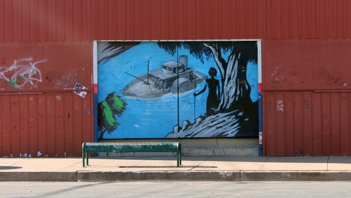 A mural on a red wall depicting a paddle steamer boat floating down a blue river watched over by aboriginal people.