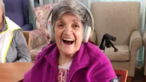 A woman with grey hair and a bright top, wearing headphones and sitting in a chair.