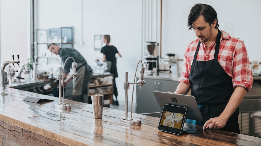 Male barista stands behind a wooden bar in a coffee shop for a story about mental ill health in the hospitality industry.