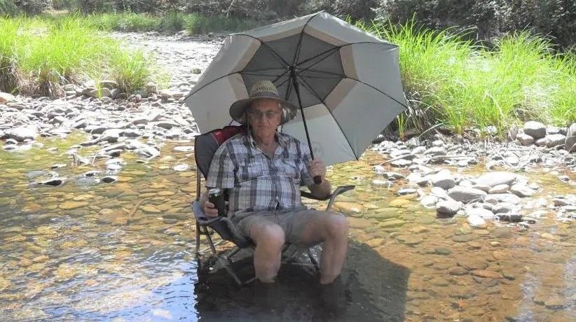 Russell Hill sits on a camp chair that is in a low creek, holding an umbrella over him, with a beer in one hand.