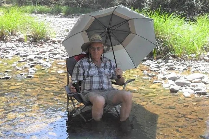 Russell Hill sits on a camp chair that is in a low creek, holding an umbrella over him, with a beer in one hand.