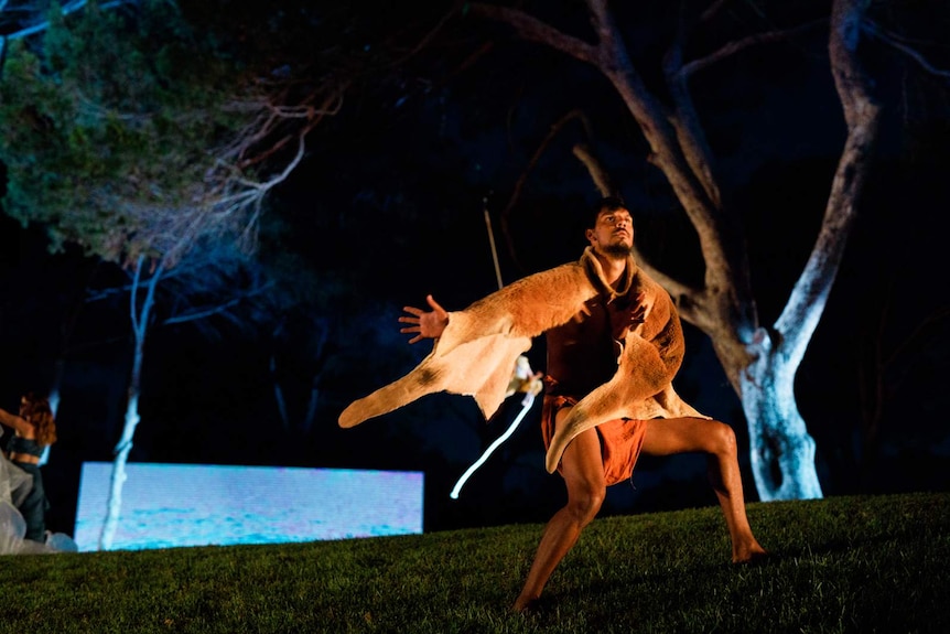 Dancer wearing skins on otherwise bare torso, outdoors, striking a pose on grass with trees and night sky behind.