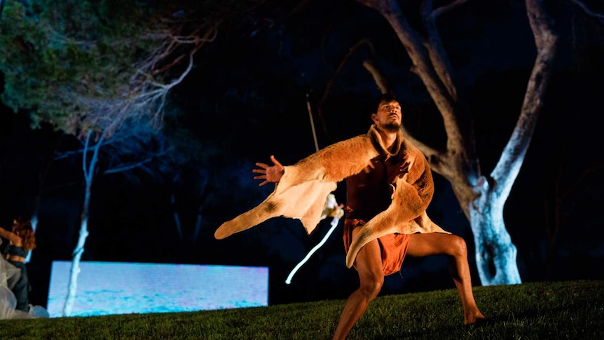 Dancer wearing skins on otherwise bare torso, outdoors, striking a pose on grass with trees and night sky behind.