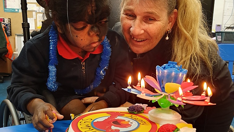 A girl in a classroom blows out candles on a birthday cake with Elmo characters on it.