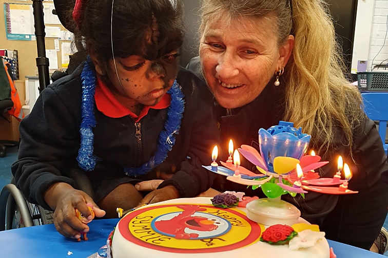 A girl in a classroom blows out candles on a birthday cake with Elmo characters on it.