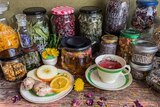 A collection of jars of dehydrated ingredients, colourful petals and leaves, arranged on a bench around a cup of ginger tea.