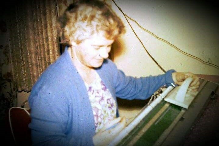 An old photograph from the 60s shows a woman in a cardigan sitting at a sewing machine, smiling.