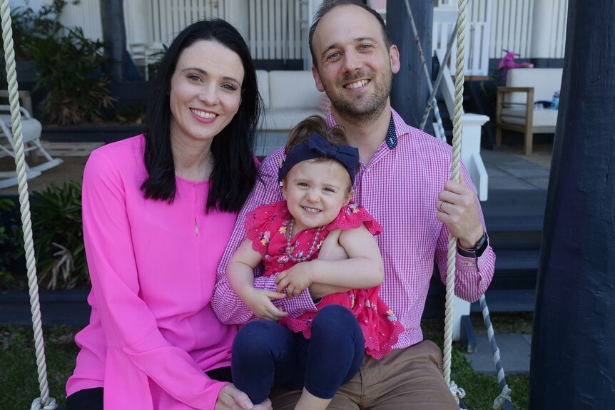 Catherine and Tim Rivett wear bright pink shirts while they sit on a swing with Charlotte on their laps.