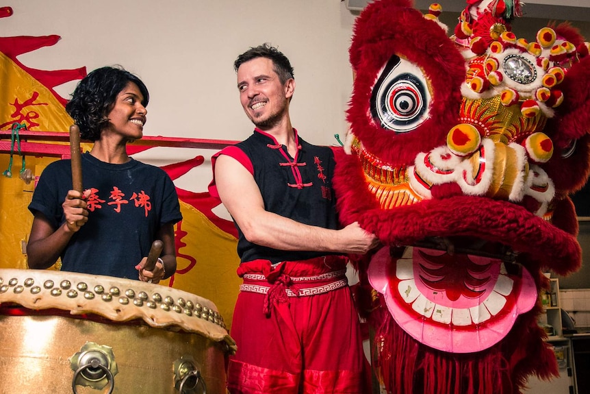 A woman posing with a drum and a man posing with a red and gold Chinese lion dancing costume head.