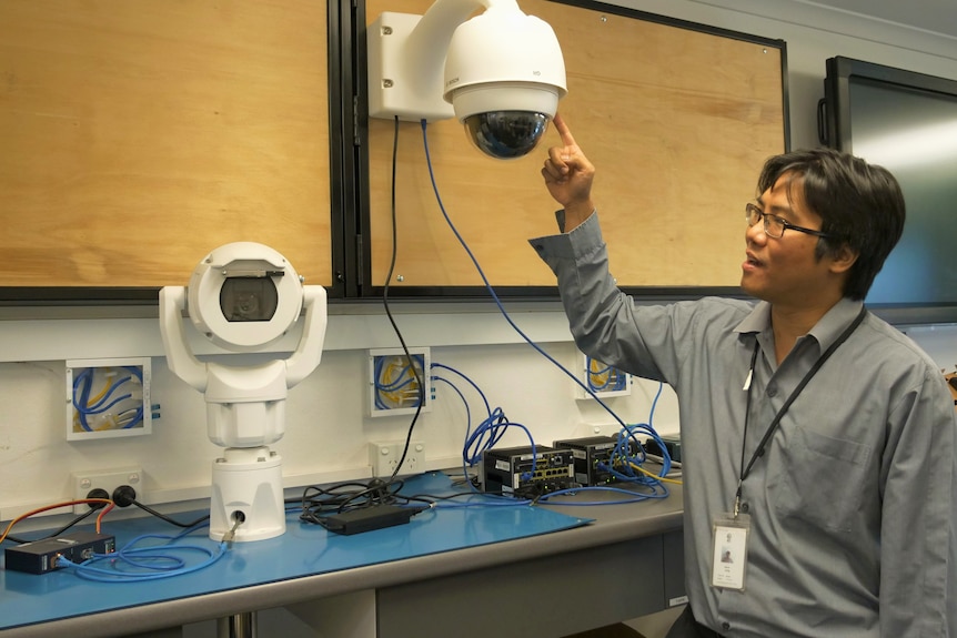 A man in a suit points to a white, round camera, used to monitor roads.