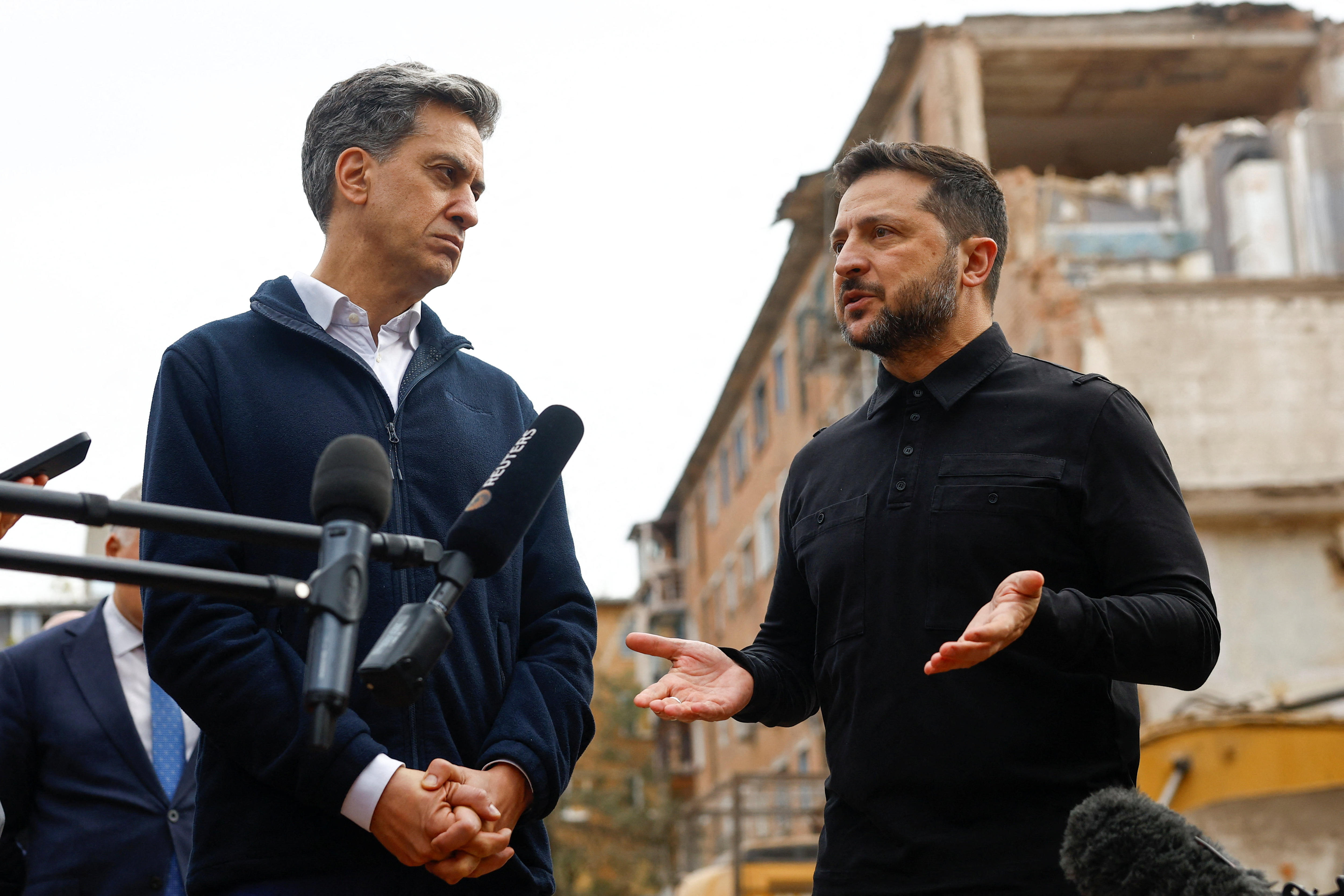 Ed Miliband (left) and Volodymyr Zelenskyy stand in front of a damaged apartment building, and speak to reporters.