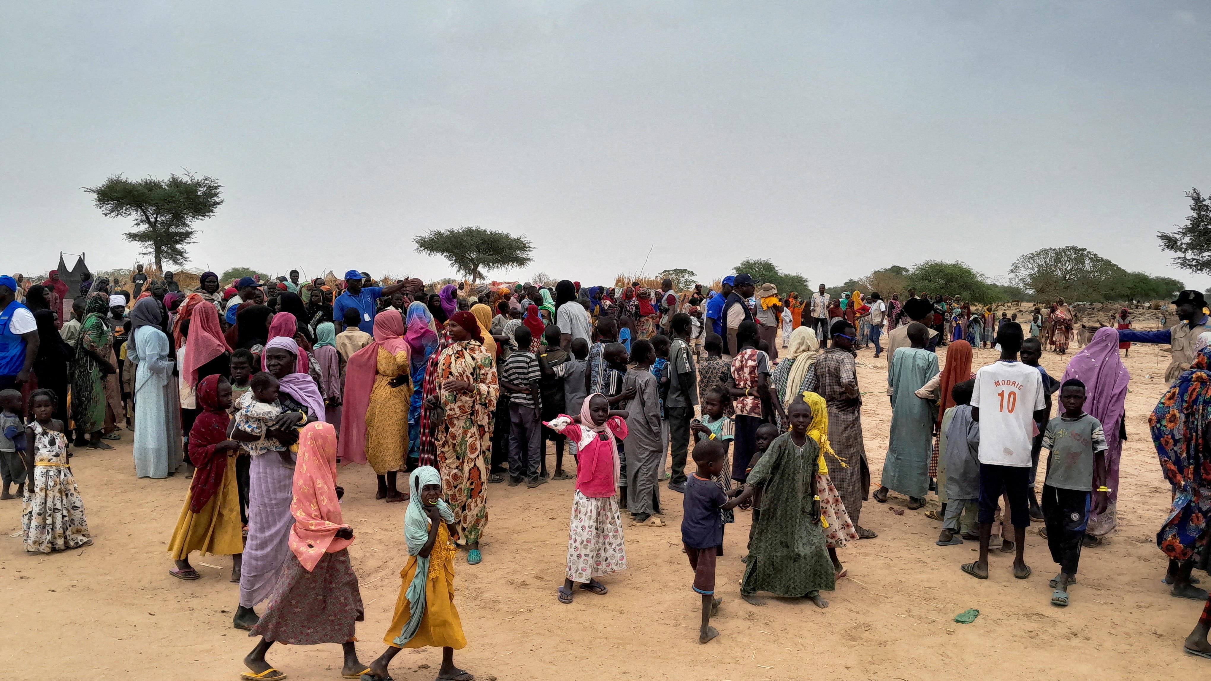 A large crowd of African women and children in colourful clothing wait in queues in a sandy desert environment.