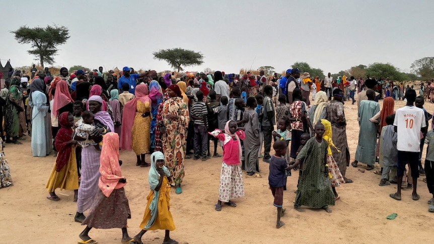 A large crowd of African women and children in colourful clothing wait in queues in a sandy desert environment.