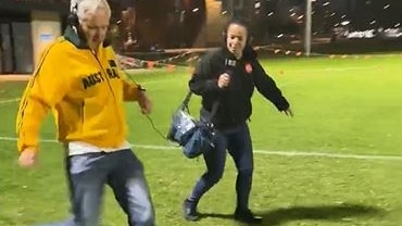 man in yellow trachsuit top about to kick a soccer ball on an oval with woman in black with headphones on in background