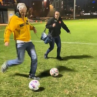 man in yellow trachsuit top about to kick a soccer ball on an oval with woman in black with headphones on in background