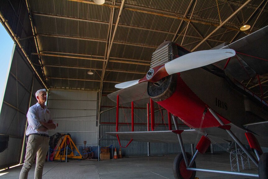 A man in a white shirt with his arms crossed stands inside an old aviation hangar and looks at a red and silver propeller plane.