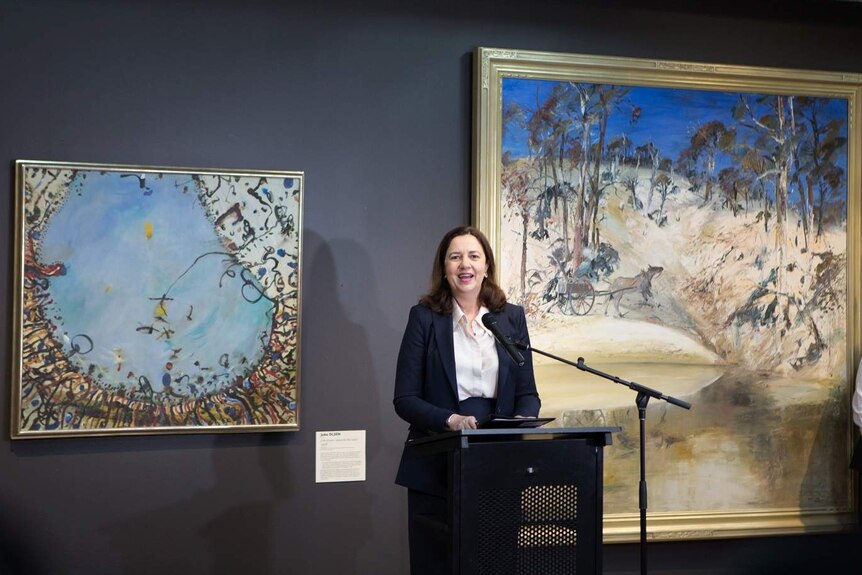 A woman with brown hair in a suit stands at a microphone in an art gallery with two large paintings hanging on the wall.