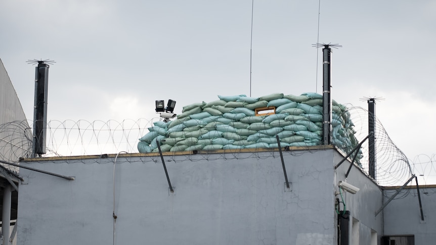 Ein Haufen Sandsäcke mit einem Loch zum Durchschießen liegt auf einem Gebäude mit Stacheldraht 