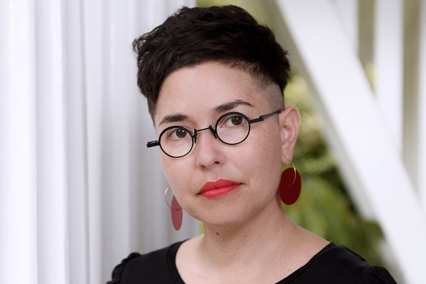 Woman with short dark hair wearing round thin-framed glasses and black tee standing next to corrugated white wall.