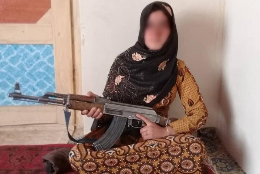 A girl wearing a head scarf and long dress sits on the floor holding an AK-47 gun.